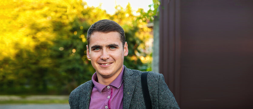 Portrait of a smiling 25-year-old young man in a tweed jacket standing outdoors against a warm green autumn background with copy space. Gen-Z workforce onboarding, quarter-life career ambition.