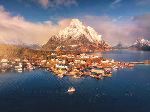 Aerial view of Reine village surrounded by fjords and snowy mountains in Lofoten, Norway. Bright winter sunlight over fishing cabins, blue sea, and dramatic coastal landscape. Top drone view