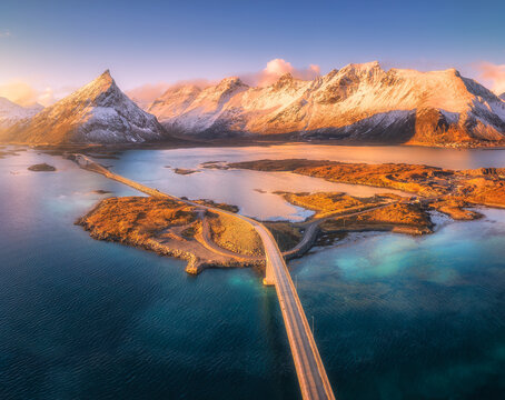 Fredvang bridges over ocean fjord with snow covered mountains in Lofoten, Norway. Aerial drone winter view of scenic road highway connecting islands at golden sunset light. Top view. Rocks in snow