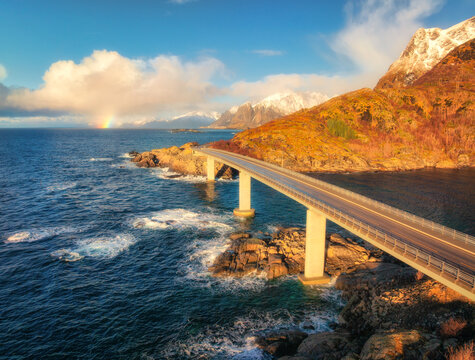 Aerial view of bridge, sea with waves and mountains at sunrise in Lofoten Islands, Norway. Landscape with beautiful road, water, rocks, blue sky with clouds and rainbow. Top view from drone