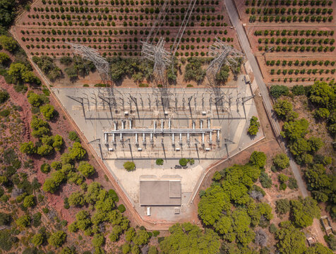 Overhead view of a triangular electrical substation with transformers and high voltage transmission towers, bordered by neat rows of citrus trees in a rural agricultural landscape