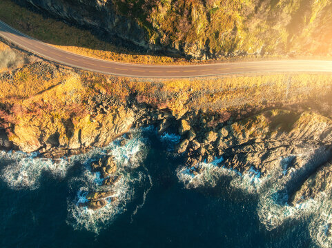 Aerial view of road, rocky sea shore with waves and stones at sunset in Lofoten Islands, Norway. Landscape with beautiful road, transparent blue water, rocks. Top drone view of highway in summer