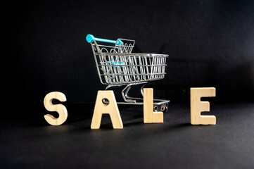 The word SALE spelled in wooden letters beside a miniature shopping cart on a dark background with colorful lighting a creative concept of shopping discounts and Black Friday deals. © Oksana