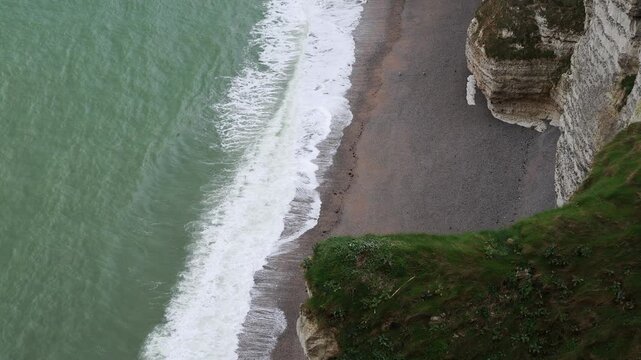 Pebble beach and breaking surf at Etretat in Normandy, France. High angle view, white foam on grey shingle, waves rolling ashore, green sea and chalk cliffs on the Alabaster Coast