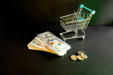 A miniature shopping cart with a black bank card beside US dollar bills and coins on a dark background a conceptual image of online shopping and card payment. © Oksana