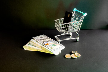 A miniature shopping cart holding a black bank card above a stack of dollar bills and coins on dark a creative concept of online shopping and card payment. © Oksana