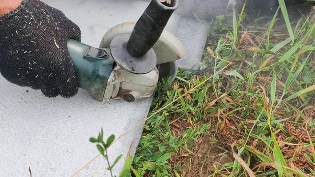 Strong worker hands in gloves hold powerful angle grinder tightly outdoors. Skilled worker hands control electric tool precisely during concrete cutting process near green grass close up.