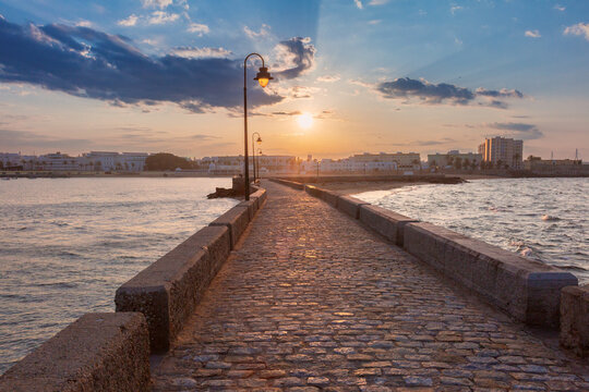 Morning sun illuminates a historic stone walkway surrounded by sea near the waterfront of Cadiz, Spain.