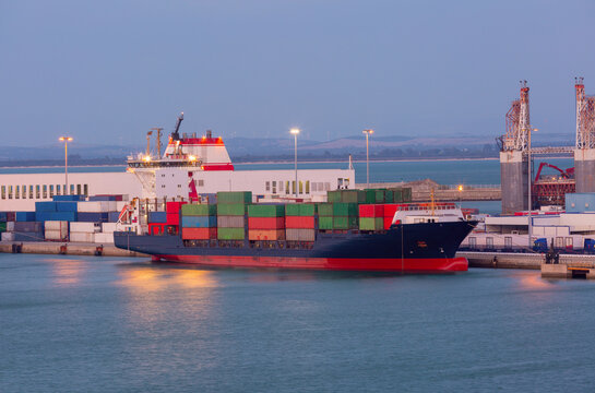 Loaded container vessel docked beside terminal facilities and port infrastructure during blue hour in Cadiz, Spain.