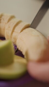 Close up of hands slicing peeled banana into thin rounds. Knife moves steadily through fruit creating incremental cuts. Green sliced object remains in foreground throughout
