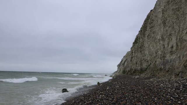 Chalk cliff and pebble beach at Cap Blanc-Nez, France. Panning shot of waves breaking on the shingle shore below a steep white rock face on the Opal Coast under a grey overcast sky