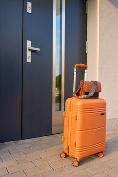 Orange rolling suitcase with handbag stands beside modern front door outdoors