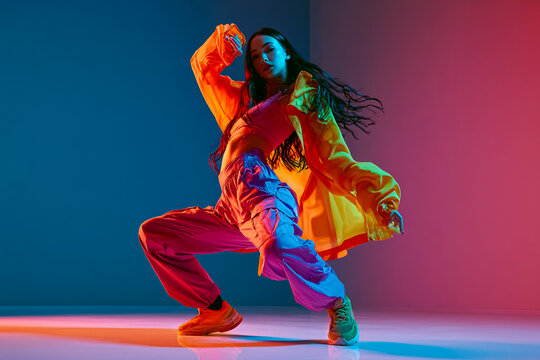 A stylish young woman performing energetic hip-hop dance moves in a studio with vibrant dual-tone neon blue and pink lighting, wearing trendy streetwear clothing, dynamic pose, modern urban lifestyle