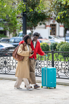 African american married couple man and woman travelers walking along street in new city, using phone. Young black male and female partners hugging talking and checking hotel direction on smartphone.