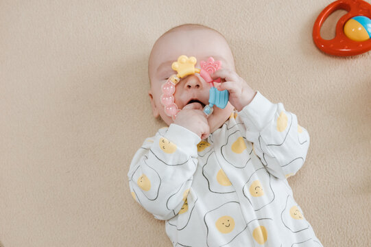 Baby lying on bed and gripping teething ring with both hands. Child chewing toy for teething relief. Exploring textures while soothing sore gums. Sensory discovery at home.