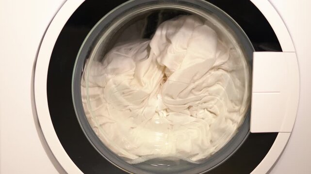 Washing machine drum spinning with laundry. Close-up of a modern front-loading washing machine during a wash cycle, with white laundry tumbling and rotating inside the spinning drum filled