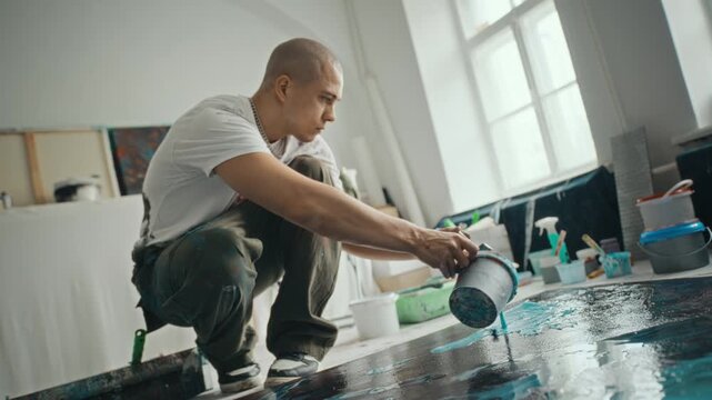 Young adult Caucasian man pouring blue paint from bucket onto large canvas on floor in art studio. Footage is suitable for creative industry marketing or art school advertisements