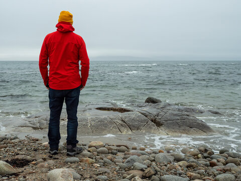 Man stands alone on rocky beach, hands on hips. Red jacket and yellow hat stand out against gray sky. Ocean waves crash, stones glisten with moisture. He gazes out, calm, thoughtful pose.