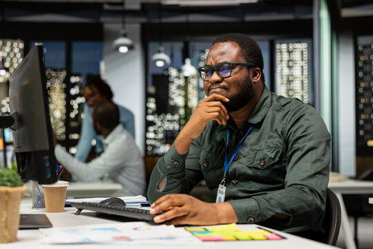 Executive assistant considers a tactical solution while rubbing his chin after hours as a black man evaluates different options through critical thinking and a different perspective at his desk.