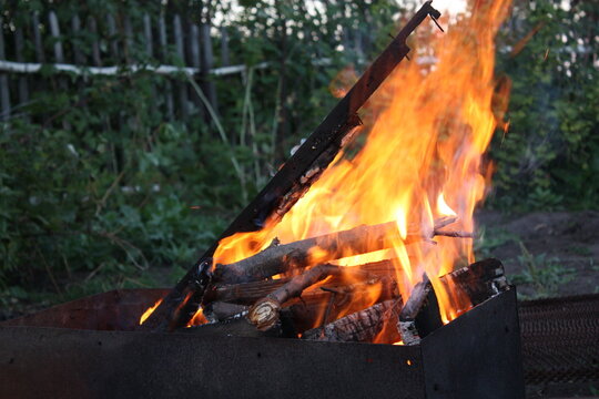 A close-up shot of hot orange flames and burning logs inside a metal barbecue grill pit in a backyard garden during evening.
