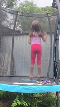 Little girl with long hair jumping on outdoor garden trampoline with safety net in backyard during summer day rear view back view concept.