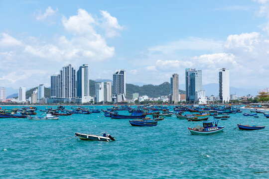 Coastal city skyline featuring modern high-rise buildings alongside a vibrant harbor filled with various fishing boats, all set against a bright blue sky with fluffy clouds