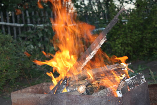 A close-up shot of vivid orange flames flickering in the wind while burning wooden logs inside a steel barbecue pit outdoors.
