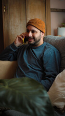 Fototapeta premium Happy young Hispanic man sitting on his sofa and talking on the phone, smiling during a cheerful conversation in a comfortable home setting.