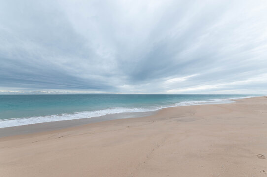 Minimalist coastal landscape with pale sand, blue water and a vast cloudy sky above the horizon