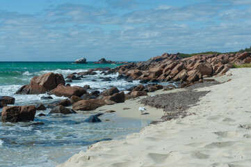 Rocky coastline with turquoise ocean, white sand and waves beneath a blue cloud-filled sky. © Anna Zaro
