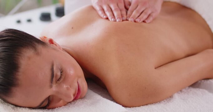 Young relaxed woman enjoying professional wellness massage in spa salon from massage therapist, lying on couch with closed eyes. Unknown skilled masseur doing manipulative treatment on back