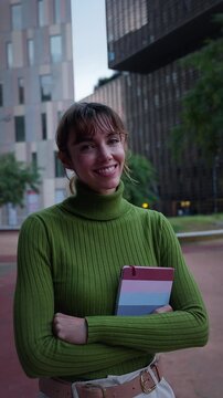 Vertical, Smiling young businesswoman standing outside a modern office looking at the camera holding a tablet arms crossed. Professional portrait conveying confidence, leadership and digital business