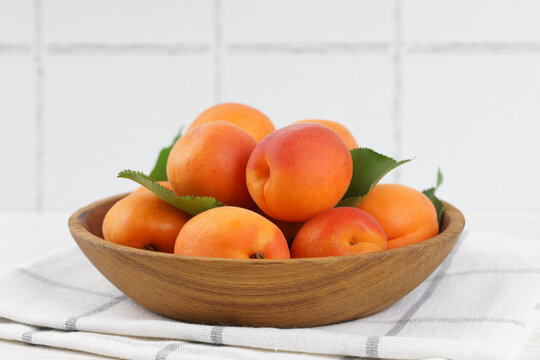 Fresh ripe apricots arranged in a wooden bowl on white kitchen table and tiled wall background with copy space. Summer stone fruits with green leaves, rustic home kitchen setting, healthy food