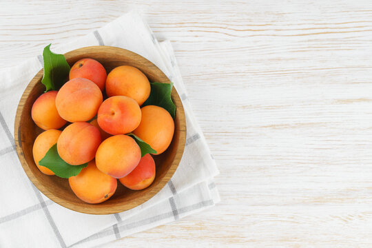 Top view of ripe apricots with green leaves arranged in wooden bowl on on white wooden background with copy space. Fresh seasonal harvest, summer fruit, healthy eating, dessert, food ingredient