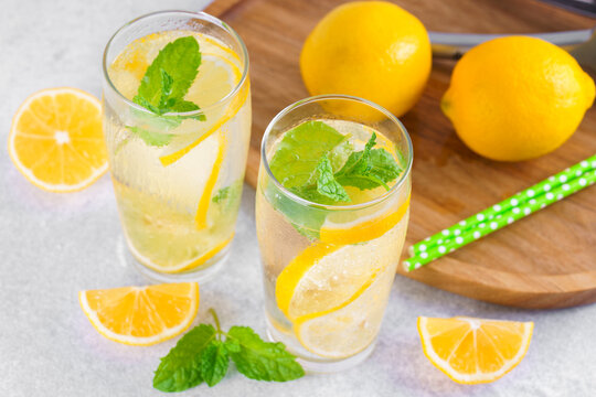 Refreshing homemade lemonade in glasses with fresh lemon slices, mint leaves, ice cubes, sparkling water served on wooden tray on white tiled kitchen background. Summer citrus beverage, drink concept.