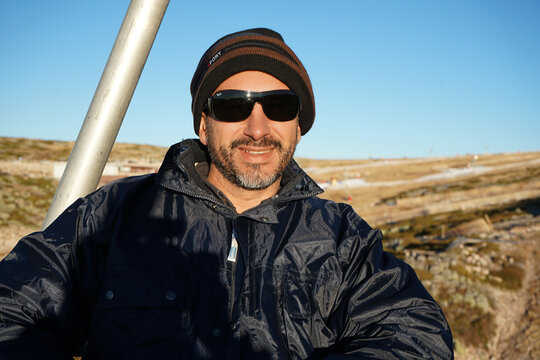 A white, dark-skinned, thin man of 40 years old, wearing a warm blue coat, a hat, and sunglasses, is on a cable car.