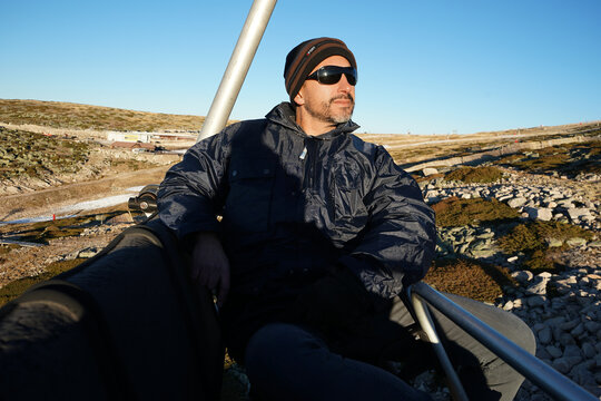 A white, dark-skinned, thin man of 40 years old, wearing a warm blue coat, a hat, and sunglasses, is on a cable car.