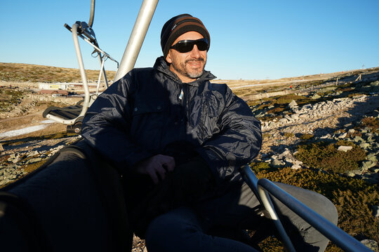 A white, dark-skinned, thin man of 40 years old, wearing a warm blue coat, a hat, and sunglasses, is on a cable car.