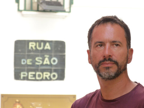 A stylish, dark-haired, thin, bearded white man of 40 years old, wearing sunglasses, with a street name sign in the background for S&atilde;o Pedro.