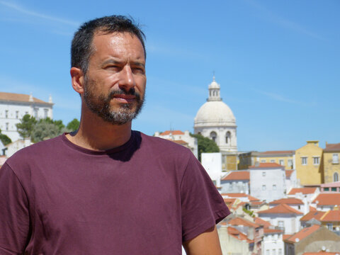 A slim, dark-haired, white European man with a trimmed beard, wearing a burgundy t-shirt, is relaxed in Lisbon, Portugal.