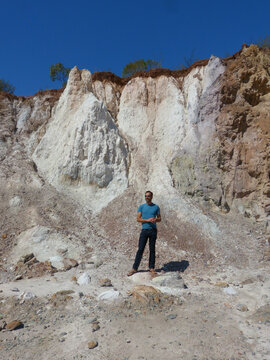 A white, dark-skinned, thin man of 40 years old, wearing trousers and a blue t-shirt, is standing near a pile of sand. A white, dark-skinned, thin man of 40 years old, wearing trousers and a blue t-sh