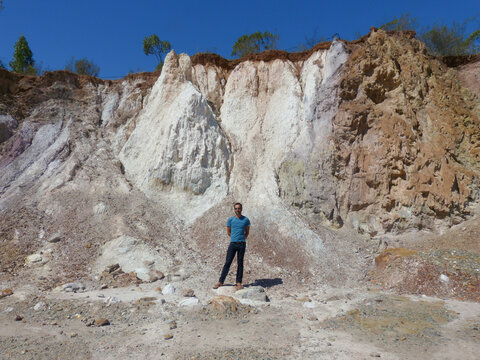 A white, dark-skinned, thin man of 40 years old, wearing trousers and a blue t-shirt, is standing near a pile of sand. A white, dark-skinned, thin man of 40 years old, wearing trousers and a blue t-sh