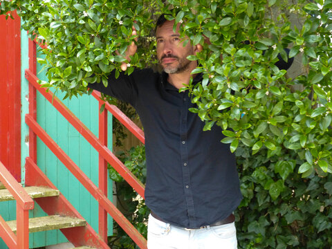 White man, dark-skinned, thin, 40 years old, casually dressed, posing against a backdrop of colorful wooden houses.