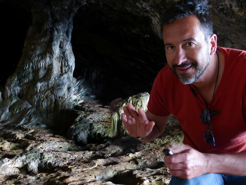 A thin, athletic-looking white man in his 40s, dressed casually, looks at the camera from inside a cave.
