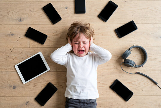 Overwhelmed young child covering ears while surrounded by smartphone, tablet and headphones, illustrating digital overload, screen addiction and technology stress.