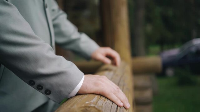 The man approaches the wooden handrail and leans on it with his hands. Close‑up shot.