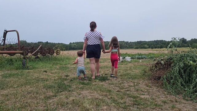 Mother walking hand in hand with her toddler boy and young daughter through a green rural field on a farm during summer day rear view.