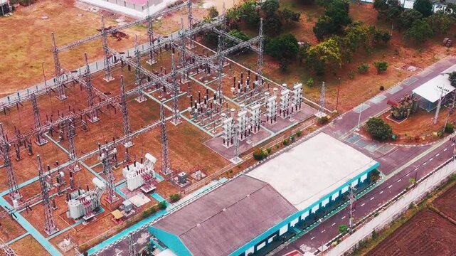 Aerial drone view of a large electrical substation featuring high voltage transformers, transmission towers, steel structures, switchgear, and power distribution equipment surrounded by dense urban bu