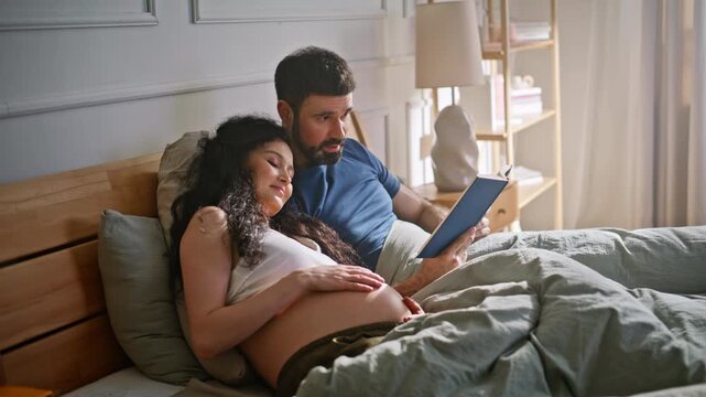 Pregnant couple reading book together bedroom. Man reads wife resting bed home.