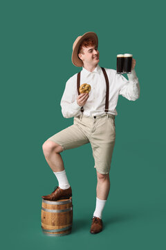 Young man in traditional German clothes with beer and pretzel on green background